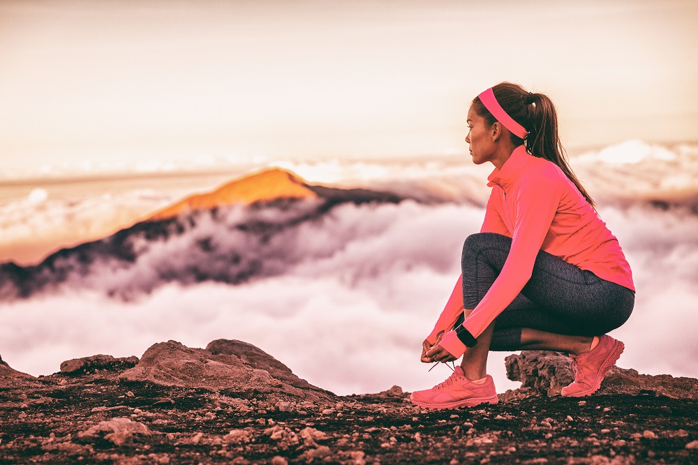 Athtlete tying laces of running shoes wearing wearable tech device smartwatch watch for fitness and sports. Trail runner woman getting ready to run on mountains nature landscape in sunset clouds.
